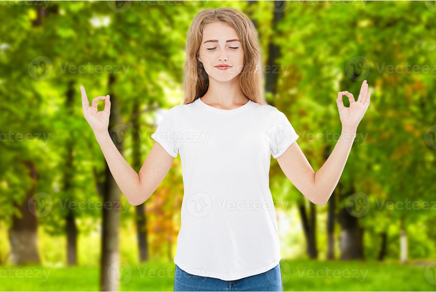 Portrait of gorgeous young woman keeping eyes closed while meditating indoors, practicing piece of mind, keeping fingers in mudra gesture on blurred park background photo