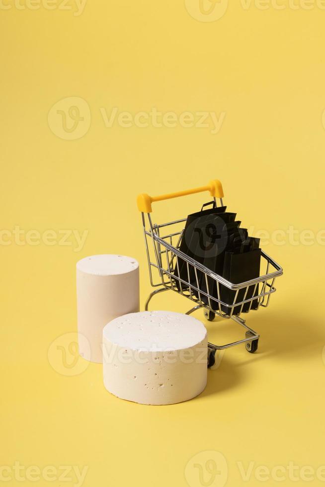 Empty mock podiums or pedestals and miniature supermarket cart with shopping bags in black friday sale on yellow background photo