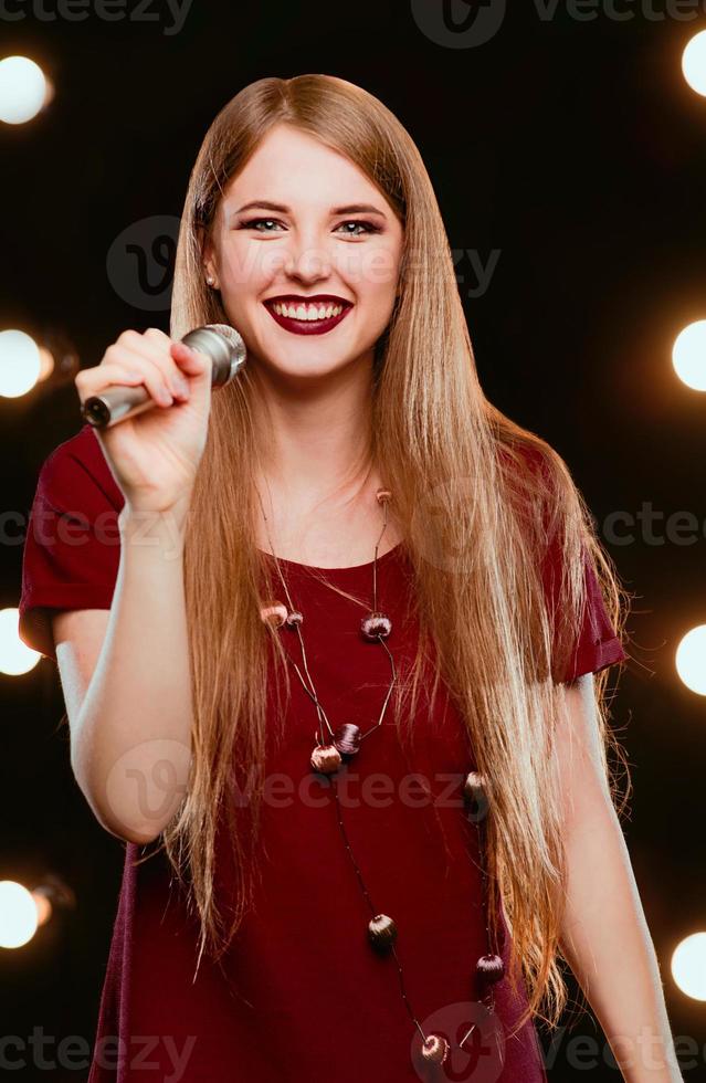 Sonriente joven hermoso cabello largo en vestido rojo mujer con micrófono cantando canciones en el escenario de karaoke foto