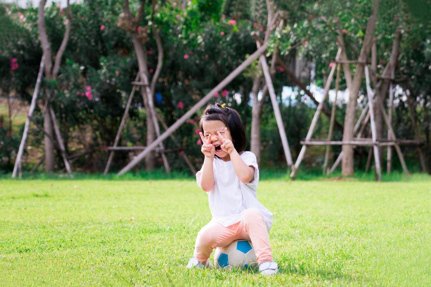 Active child girl sitting on football. Sweet smile kid. Happy children acting two finger up V-shaped. On green grass. Baby aged 4-5 years old. photo