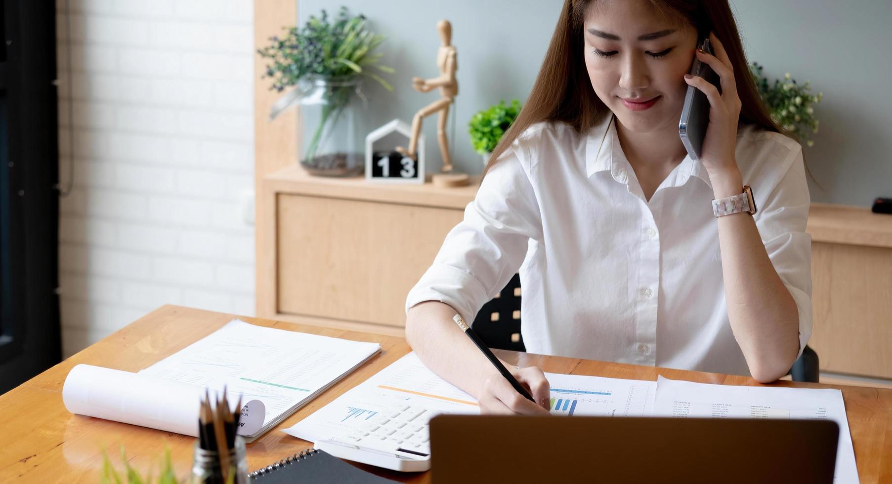 Sonriente mujer asiática que toma una llamada en el teléfono móvil durante el trabajo en una computadora portátil para obtener el número de contabilidad financiera, informe mensual de estadísticas de trámites foto