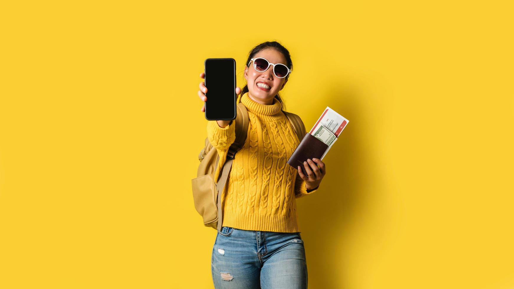 Woman traveler with suitcase, holding passport and ticket in the hand, and submit mobile phones coming in front of the camera on yellow background. Travel backpack photo