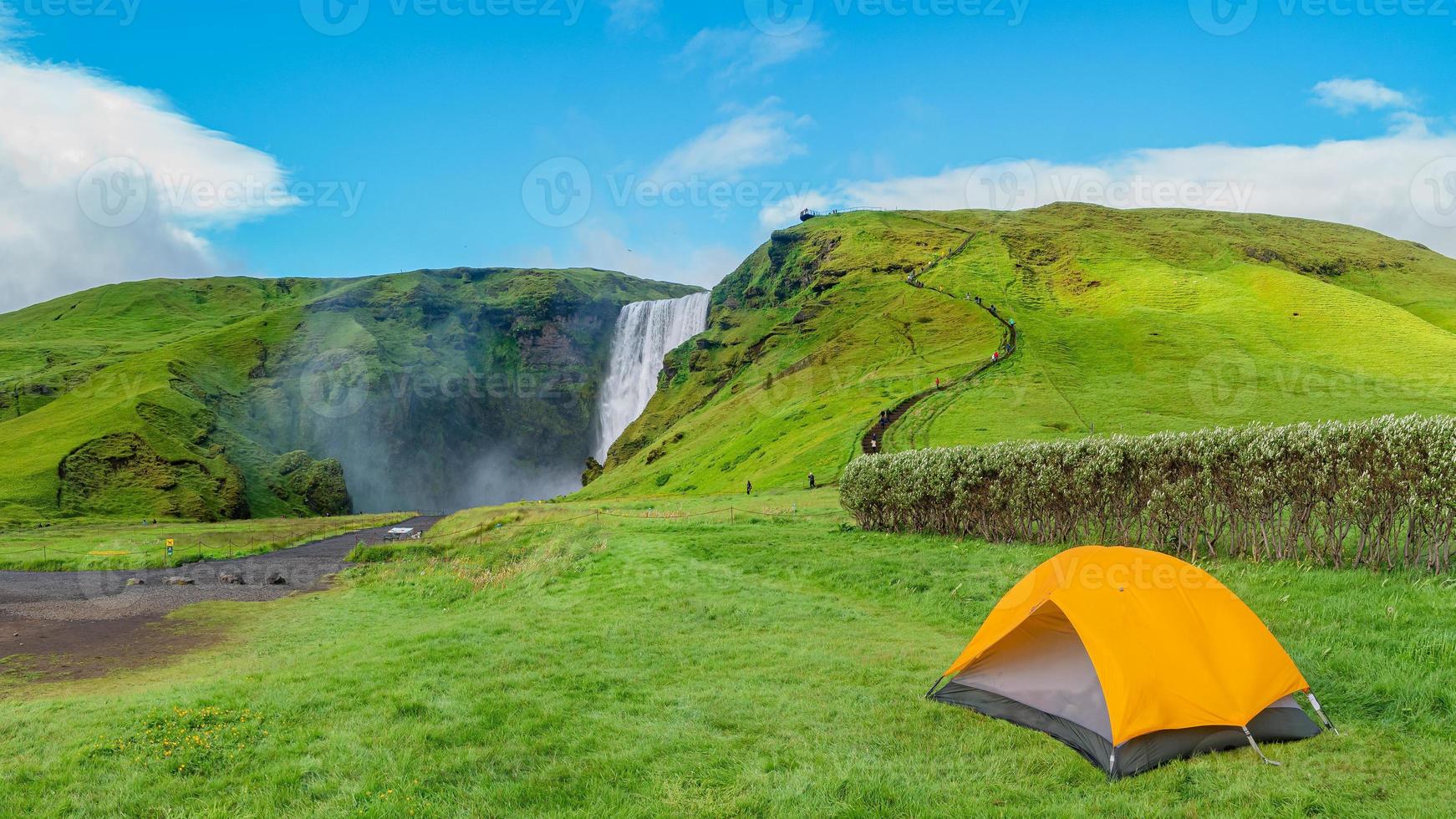 Panoramic view over camping site with orange tent, and tourists in front of famous Skogafoss waterfall, while hiking in Iceland, summer, scenic view. photo