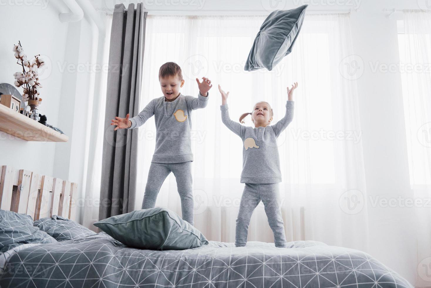 Happy kids playing in white bedroom. Little boy and girl, brother and sister play on the bed wearing pajamas. Nightwear and bedding for baby and toddler. Family at home photo