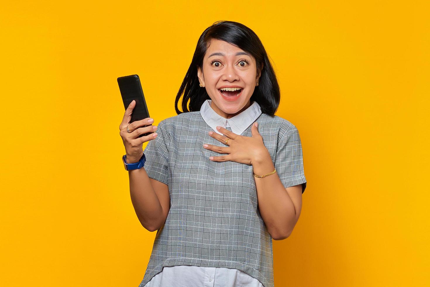 Alegre joven mujer asiática sosteniendo el teléfono móvil y poniendo las palmas en el pecho sobre fondo amarillo foto