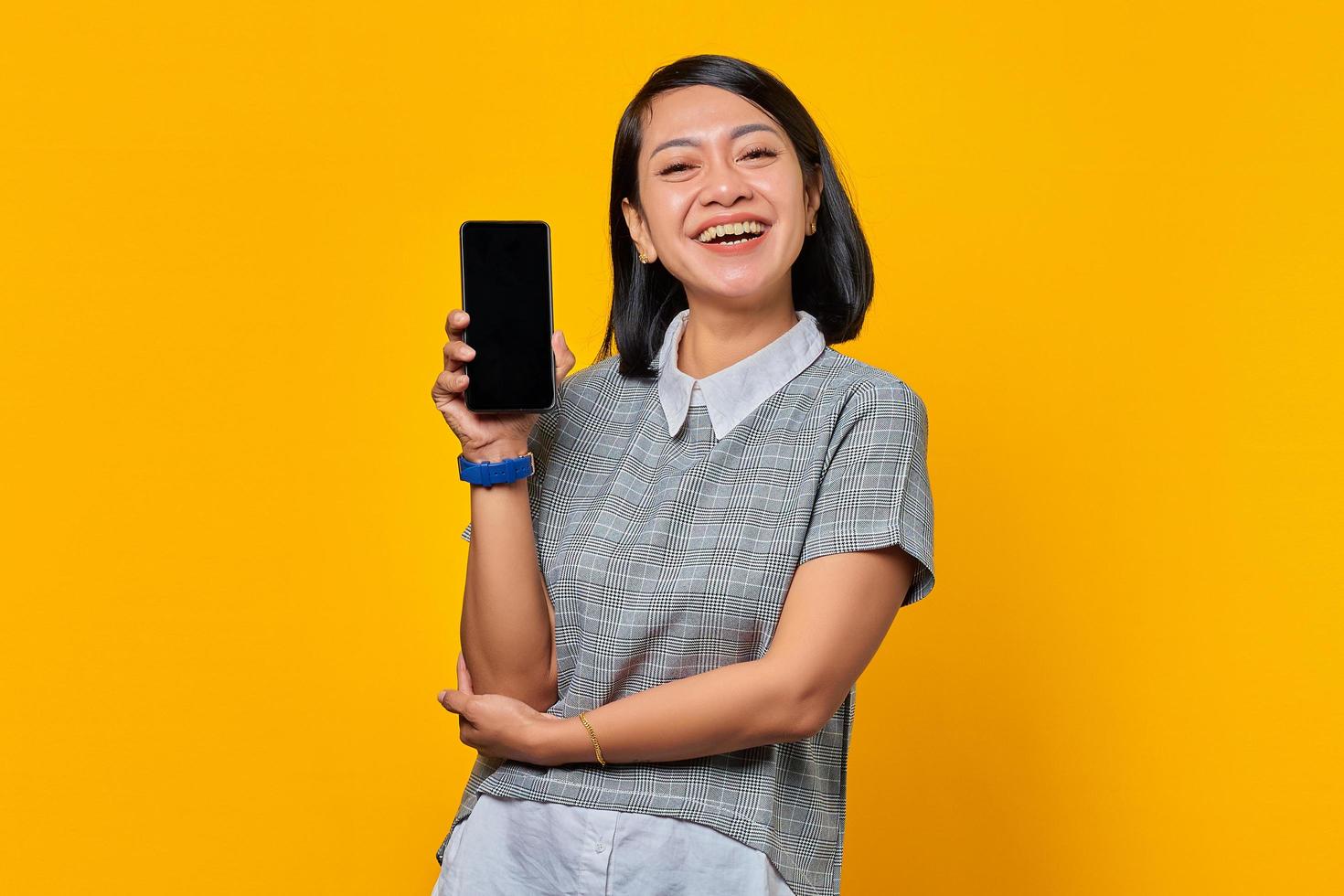 Alegre mujer asiática mostrando la pantalla en blanco del smartphone sobre fondo amarillo foto