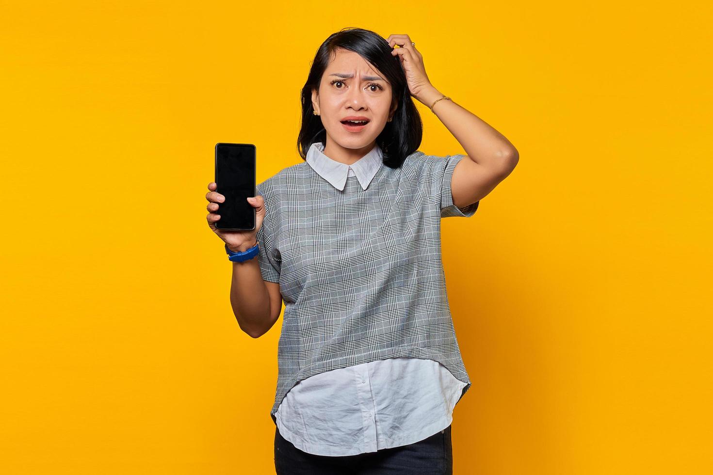 Portrait of beautiful asian woman holding smartphone touching her head and expressing concern on yellow background photo