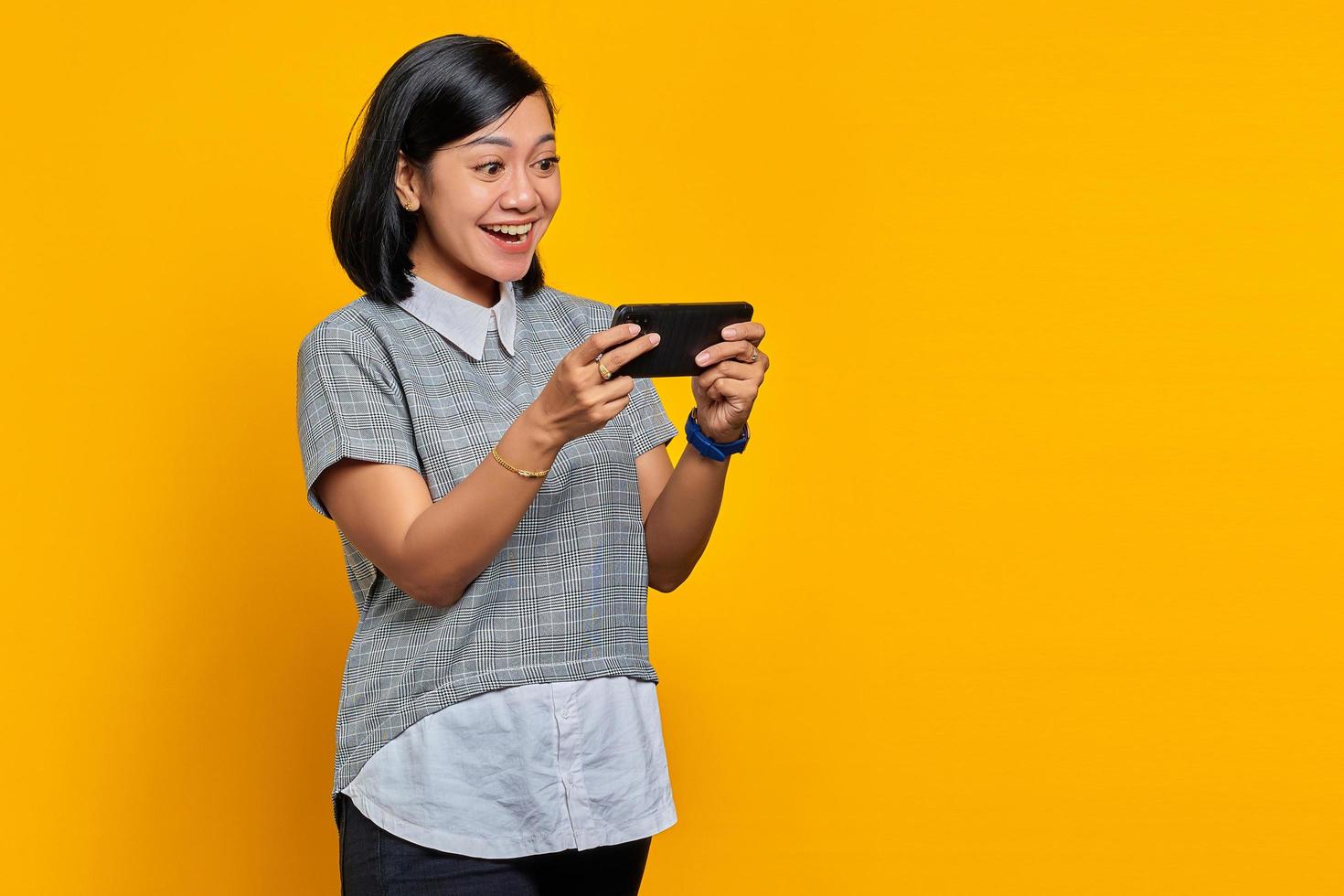 Retrato de joven alegre mujer asiática jugando videojuegos en el teléfono móvil sobre fondo amarillo foto