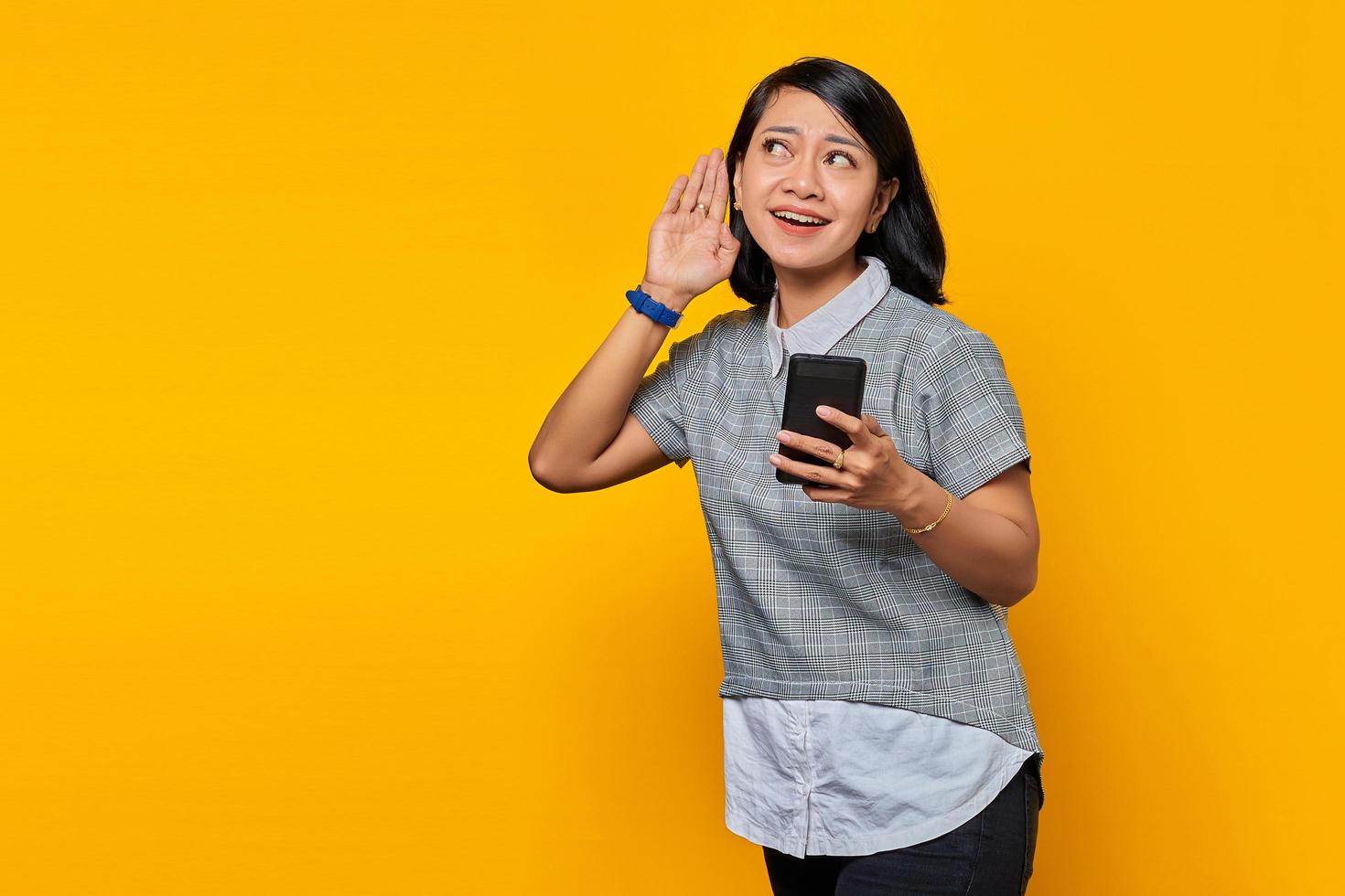 Portrait of serious looking asian woman trying to overhear secret conversation and holding smartphone over yellow background photo