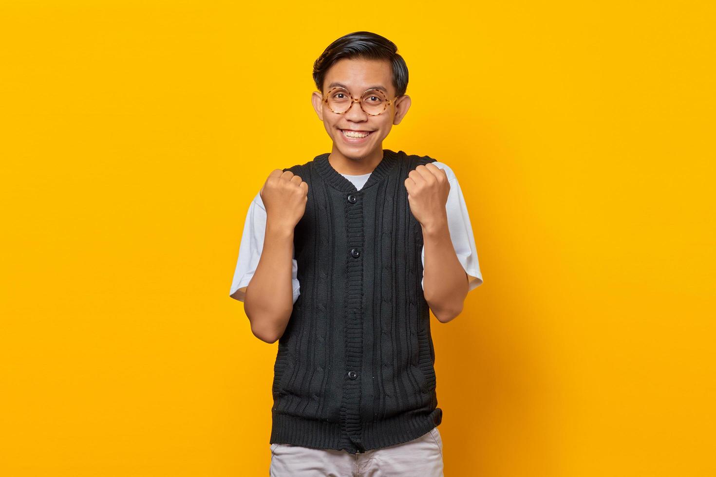 Portrait of excited young Asian man celebrating success with raised arms on yellow background photo