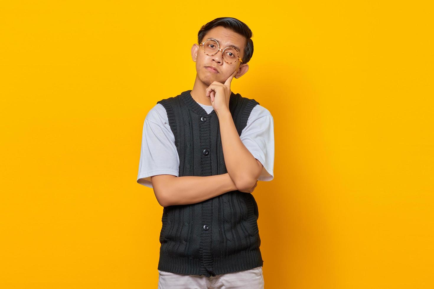 Portrait of thinking Asian man touching chin and looking at camera over yellow background photo
