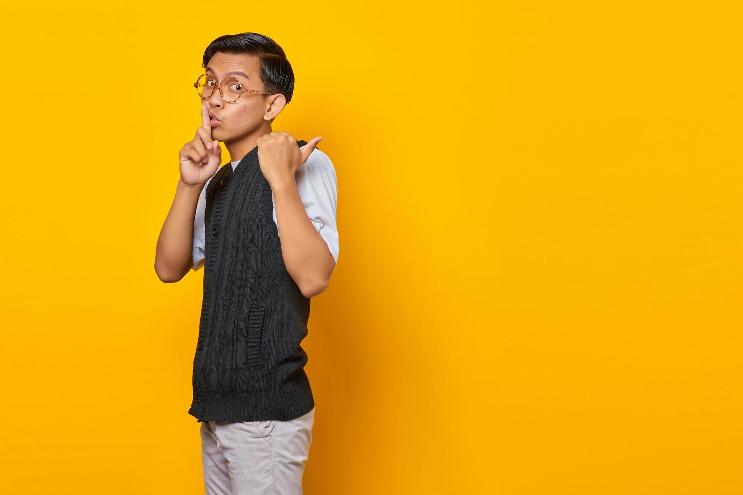 Handsome asian man making finger on lips, silent gesture and finger pointing at empty space over yellow background photo