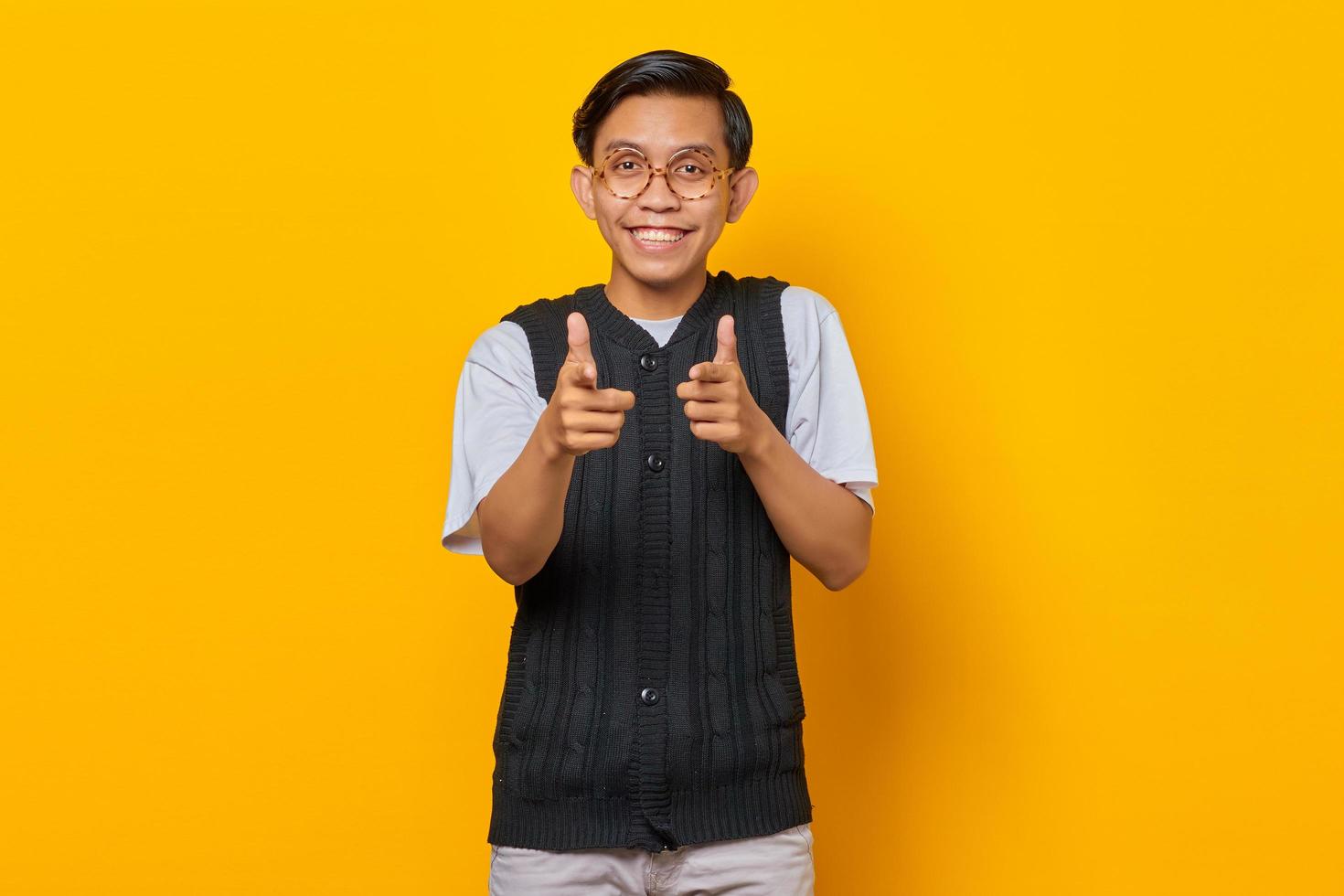 Portrait of smiling young Asian man pointing fingers at camera on yellow background photo