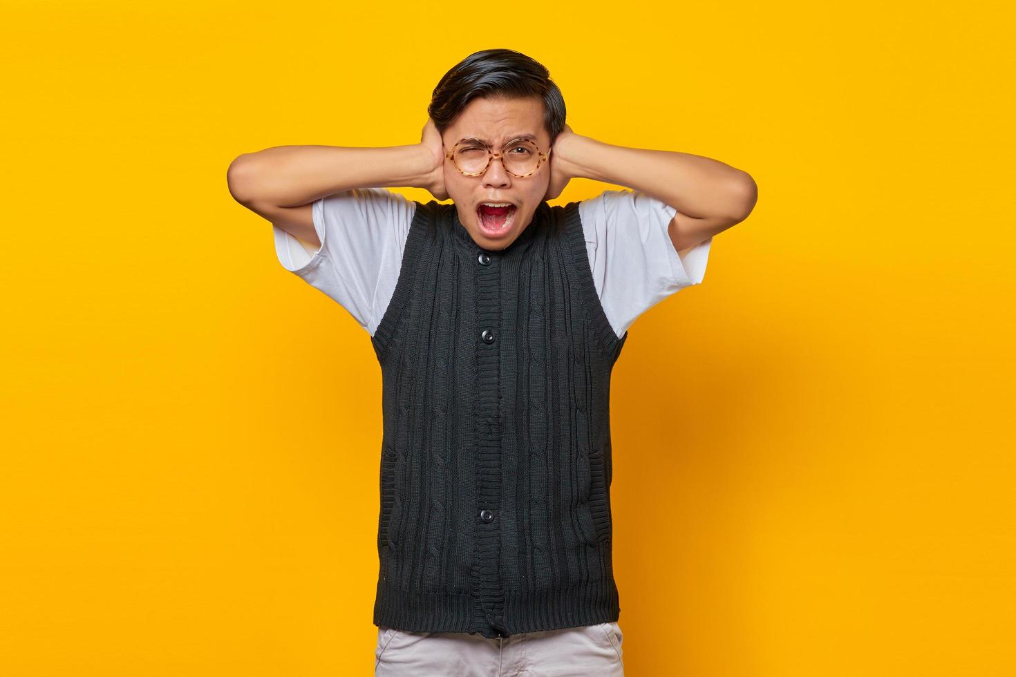 Frustrated Asian young man covering ears with hands isolated over yellow background photo