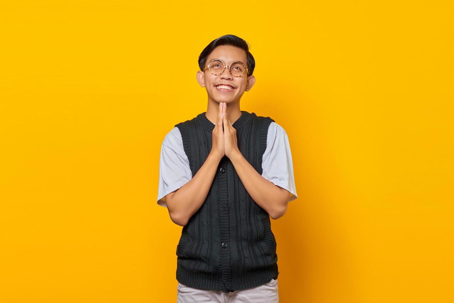 Portrait of smiling Asian young man showing prayer gesture on yellow background photo