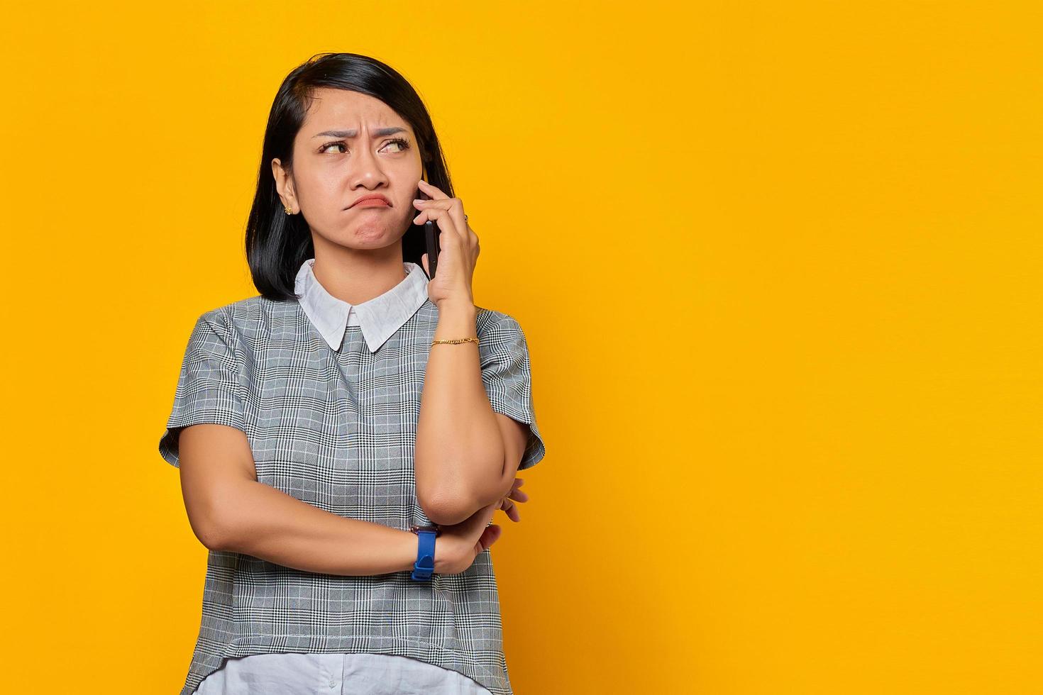 Retrato de infeliz joven asiática mientras recibe una llamada entrante en el teléfono inteligente sobre fondo amarillo foto