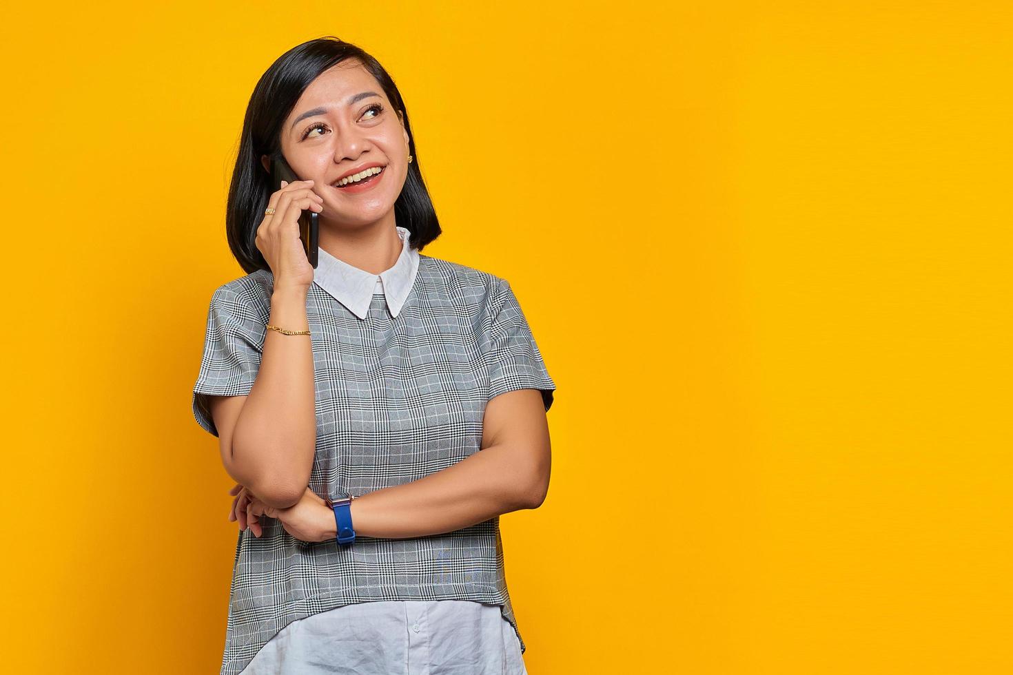 Beautiful young Asian woman receiving incoming call on smartphone and looking aside on yellow background photo