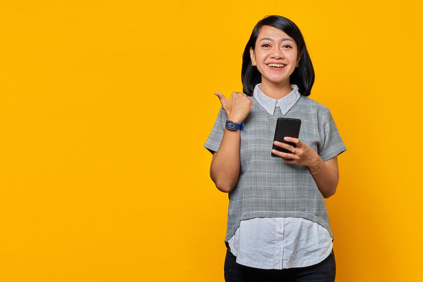 Cheerful young Asian woman holding mobile phone and pointing fingers aside at copy space isolated over yellow background photo