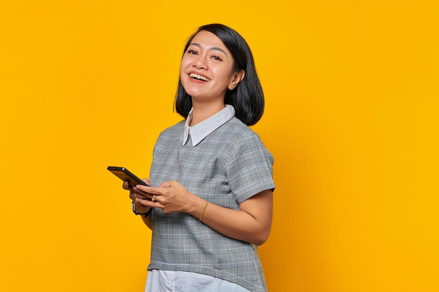 Portrait of cheerful young Asian woman holding smartphone while looking at camera on yellow background photo