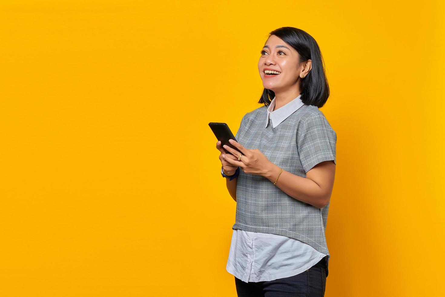 Portrait of cheerful young Asian woman holding mobile phone and looking aside isolated on yellow background photo