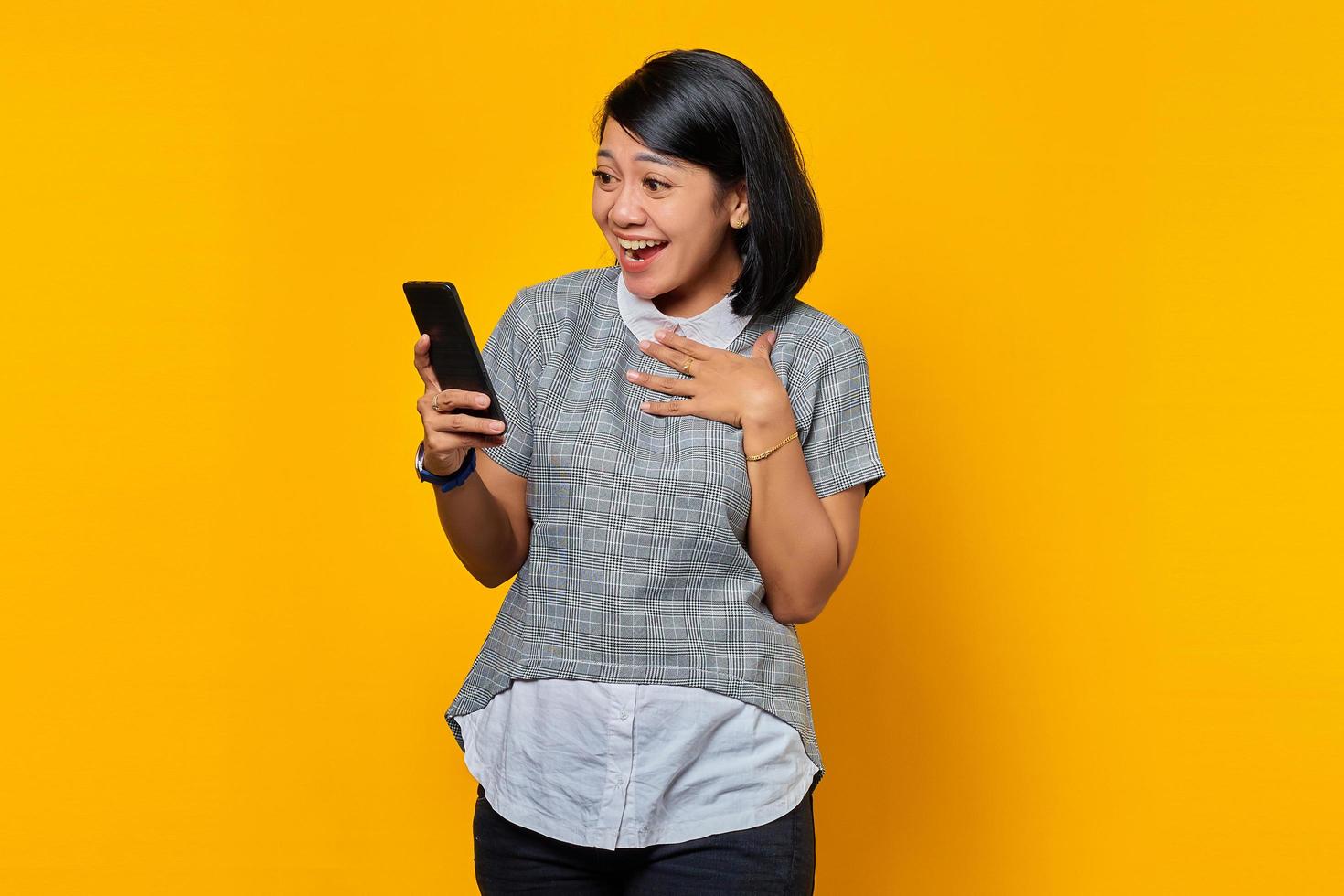 Mujer asiática joven sorprendida que sostiene el teléfono móvil con la boca abierta sobre fondo amarillo foto