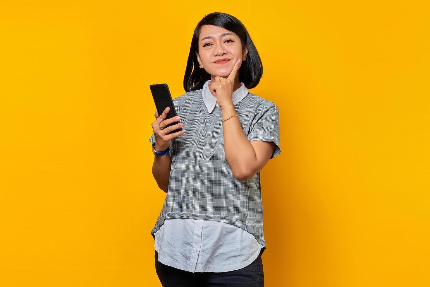 retrato, de, sonriente, joven, mujer asiática, pensamiento, pregunta, con, dedo, mano, en, barbilla, y, tenencia, teléfono móvil foto