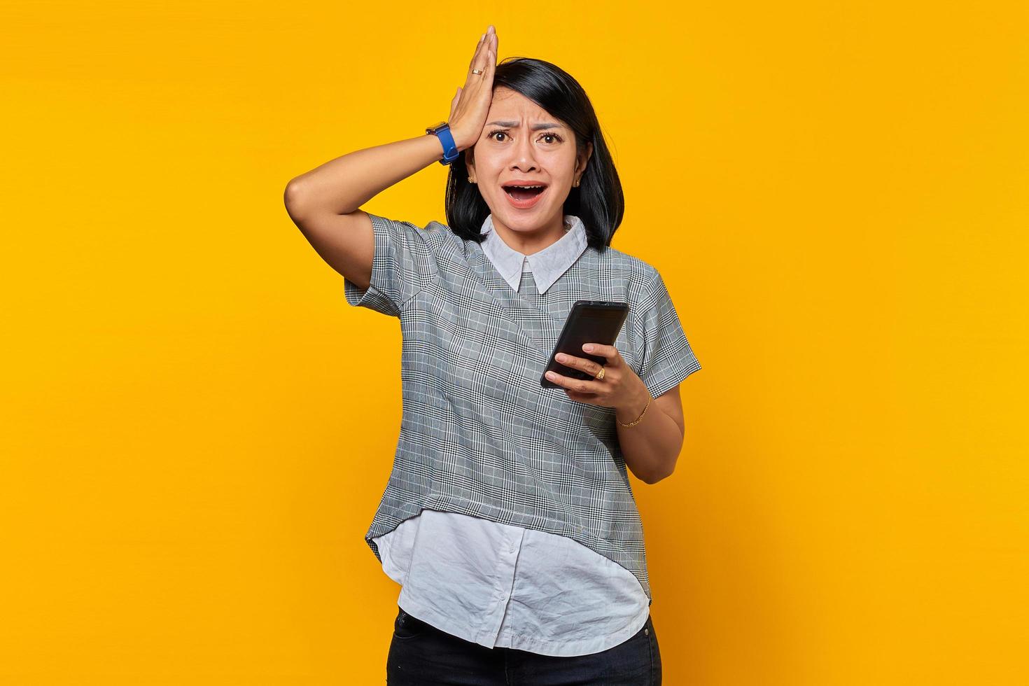 Retrato de mujer asiática joven sorprendida con la mano en la cabeza con expresión decepcionada y sosteniendo el teléfono inteligente sobre fondo amarillo foto