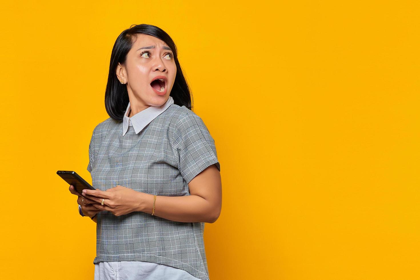 retrato, de, sorprendido, joven, mujer asiática, tenencia, teléfono móvil, y, mirar a un lado, en, fondo amarillo foto