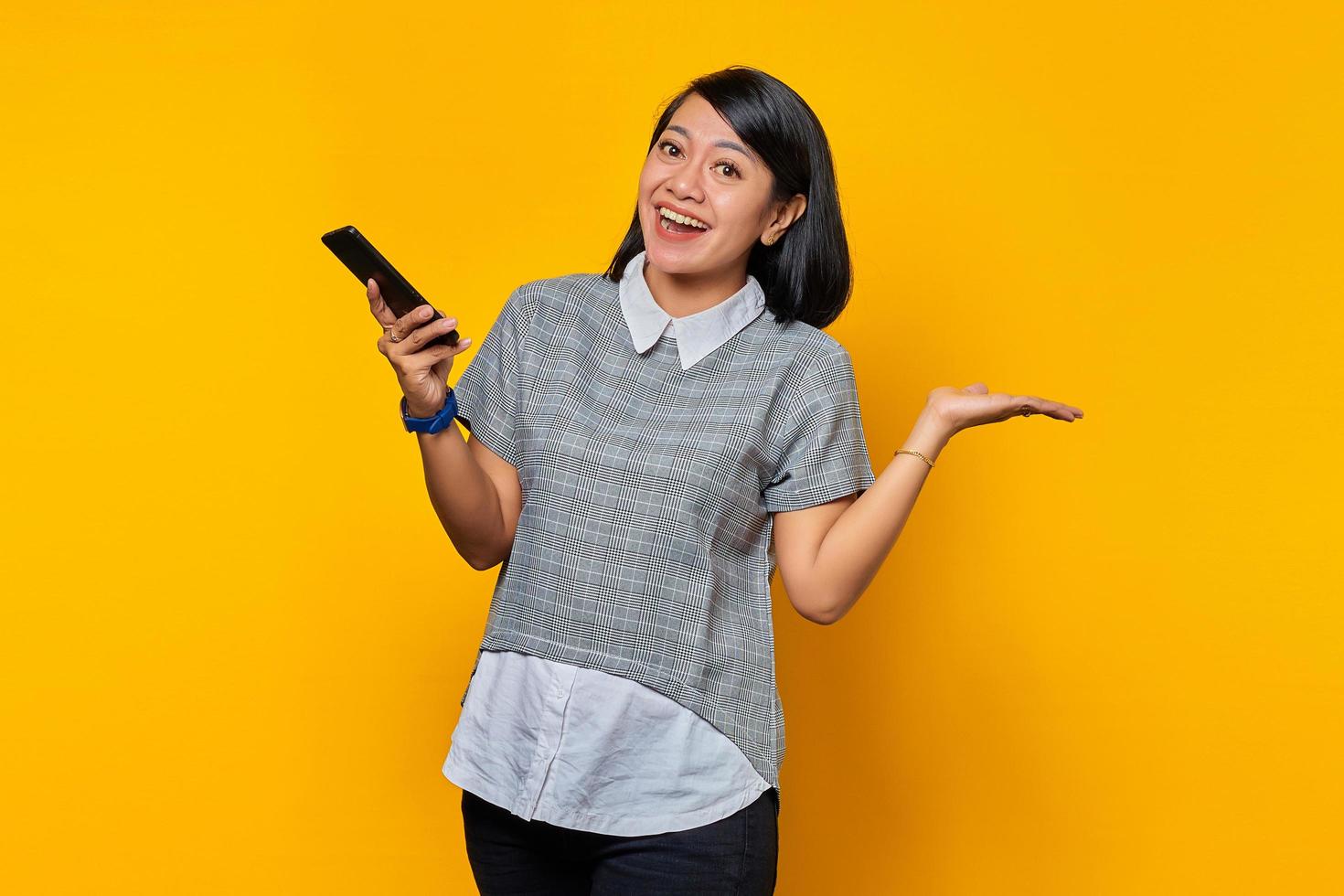 Retrato de alegre joven mujer asiática sosteniendo smartphone apuntando al espacio vacío con palmas sobre fondo amarillo foto