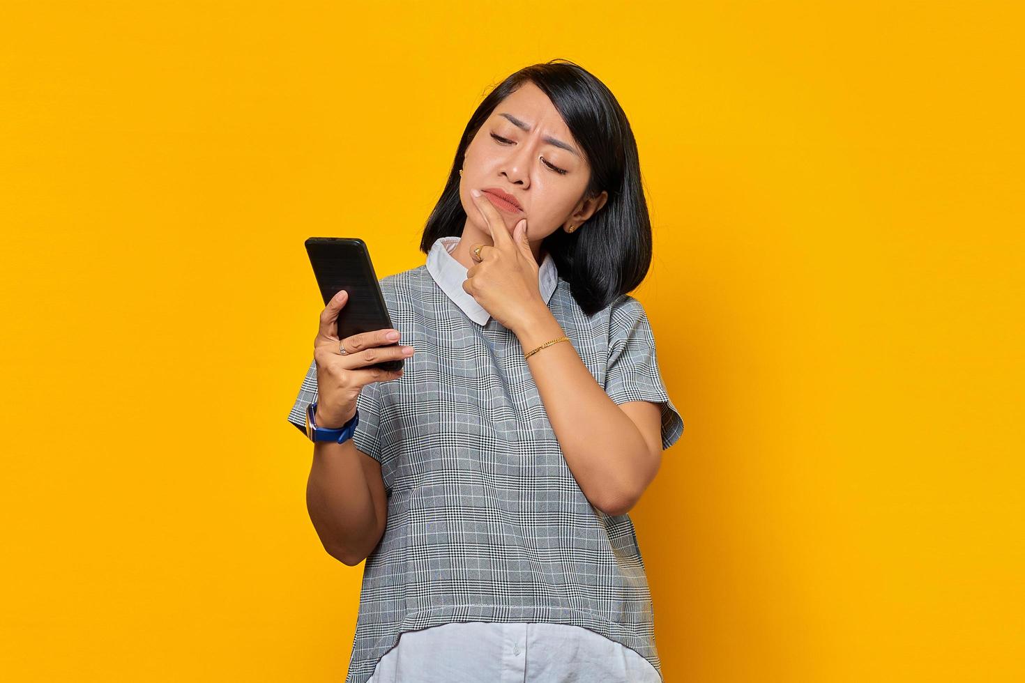 Pensativo joven mujer asiática mirando el teléfono celular con el dedo en la barbilla sobre fondo amarillo foto