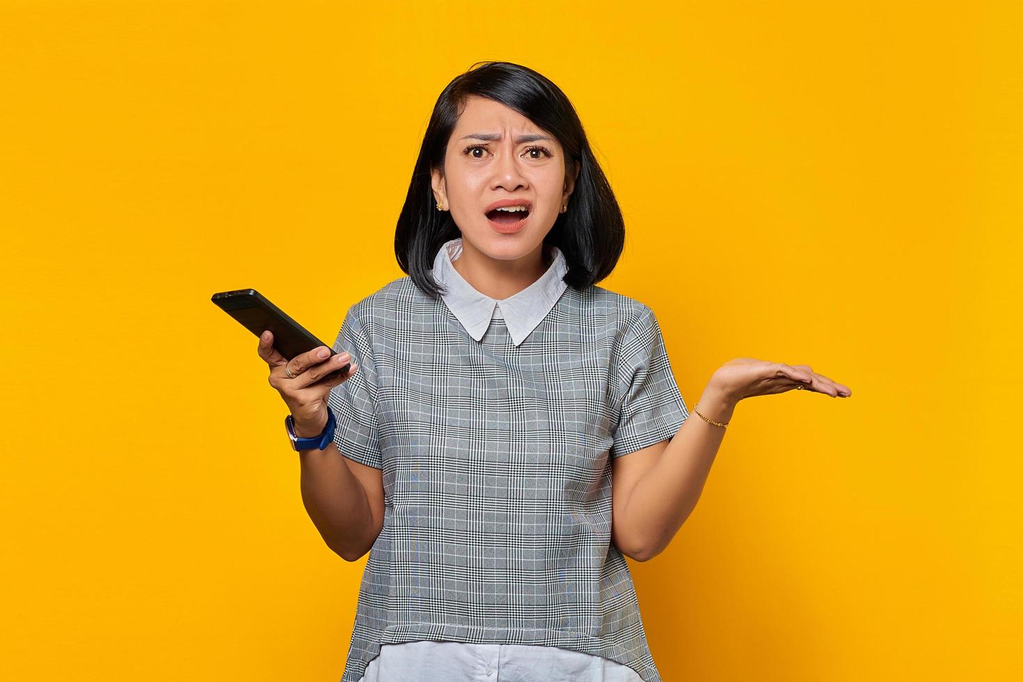 Retrato de mujer asiática sorprendida que sostiene el teléfono móvil con expresión confusa e infeliz sobre fondo amarillo foto