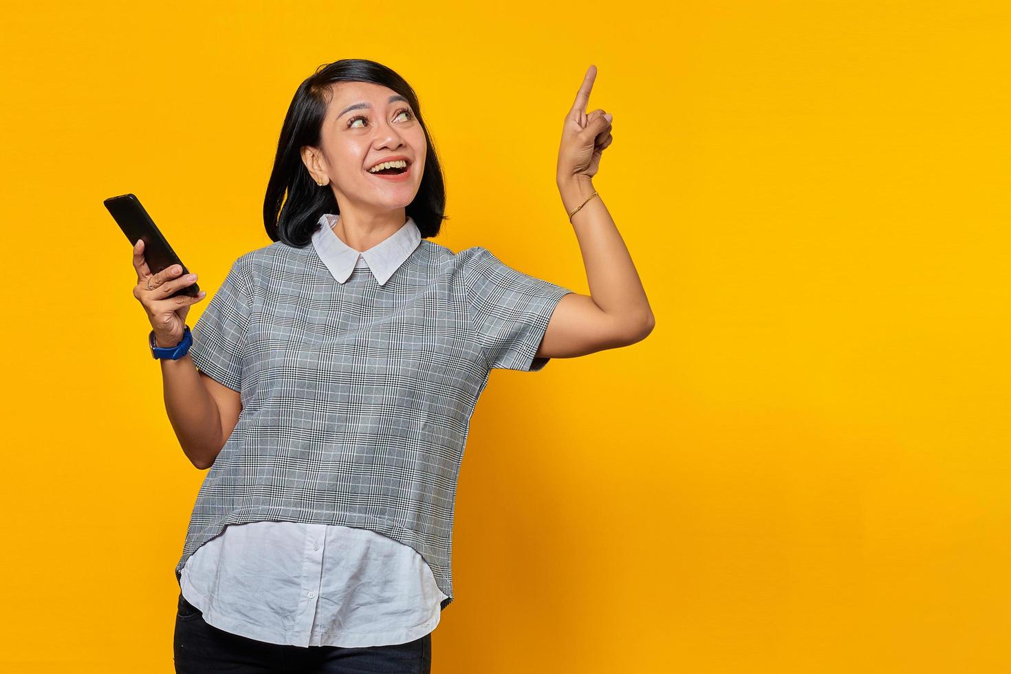 Alegre joven asiática sosteniendo el teléfono móvil y apuntando con el dedo hacia arriba sobre fondo amarillo foto