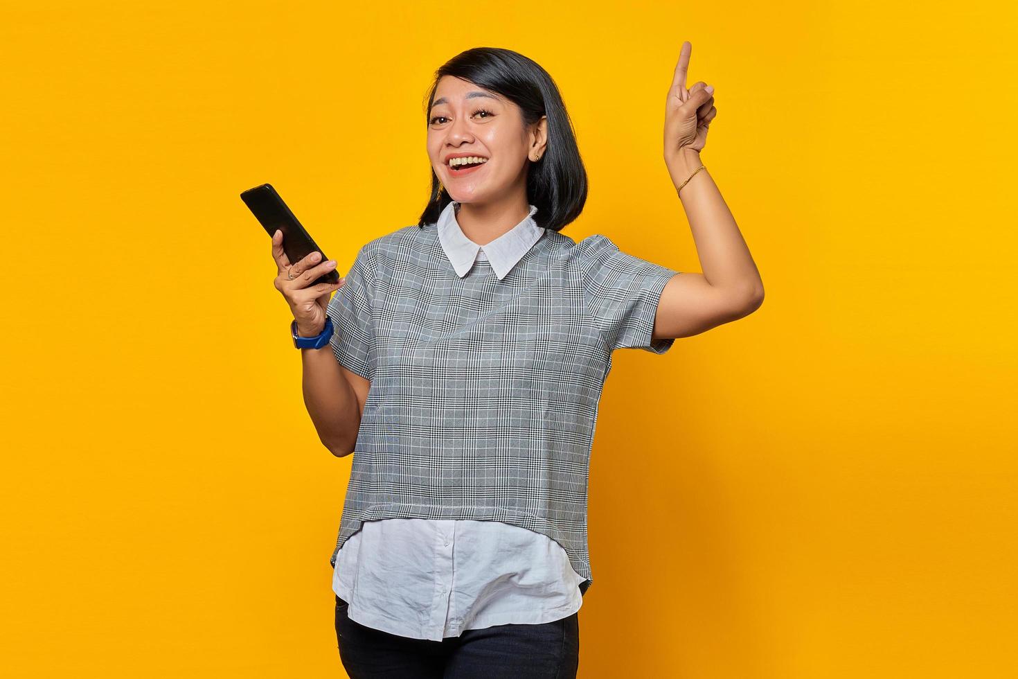 Happy young Asian woman holding mobile phone and pointing finger to copy space on yellow background photo