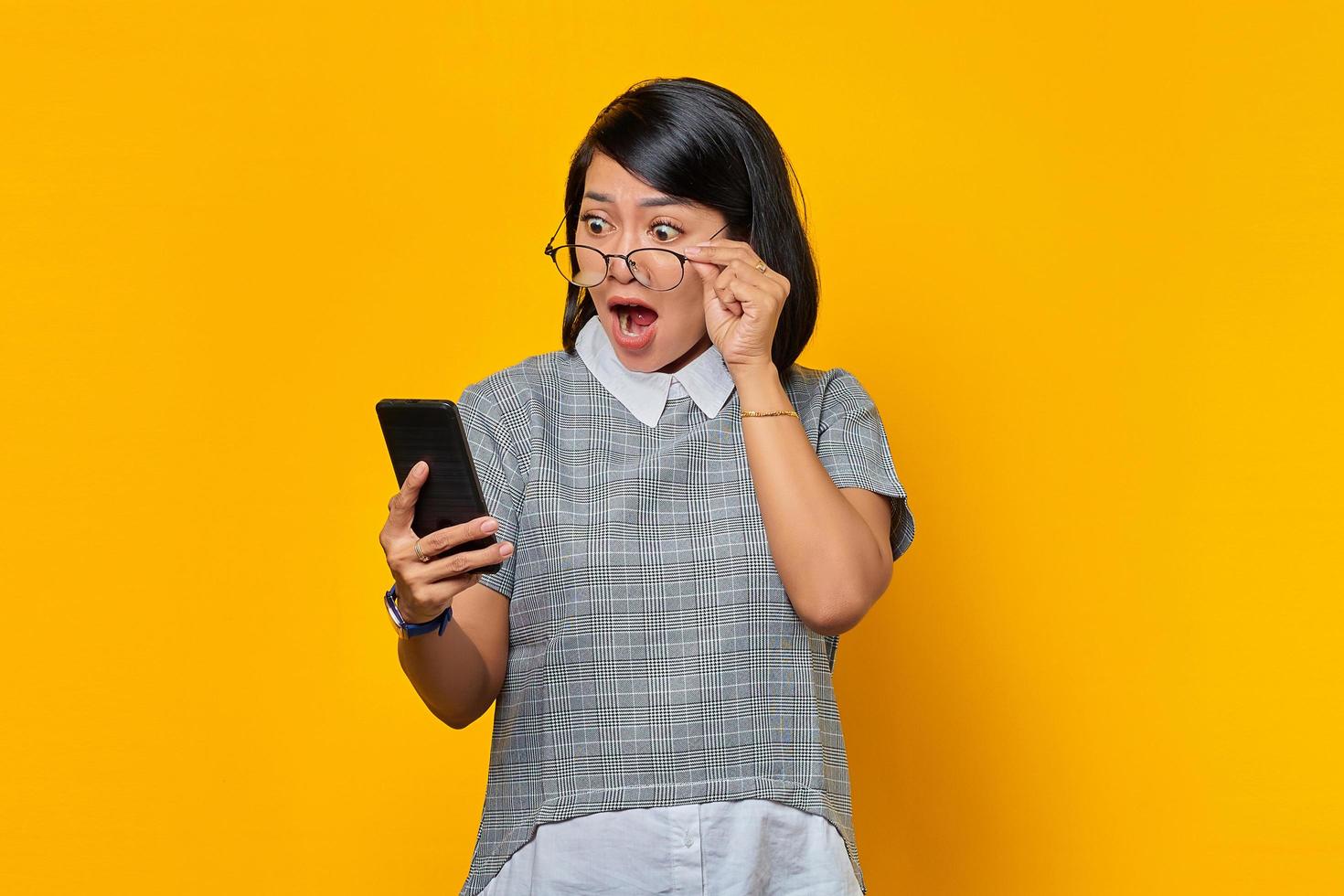 Shocked young Asian woman using mobile phone and taking of glasses isolated over yellow background photo