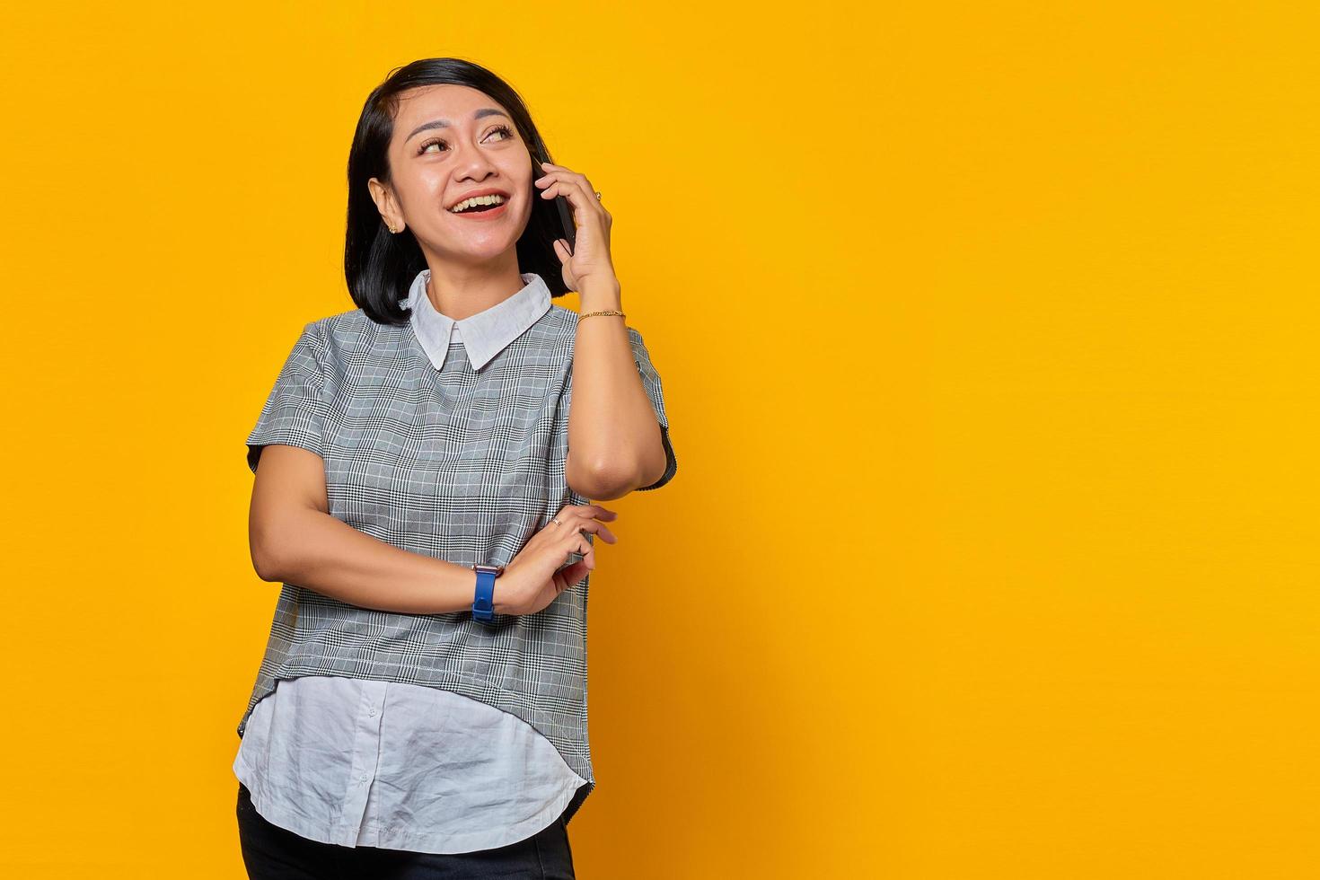 Portrait of smiling young Asian woman receiving incoming call on smartphone and looking aside on yellow background photo