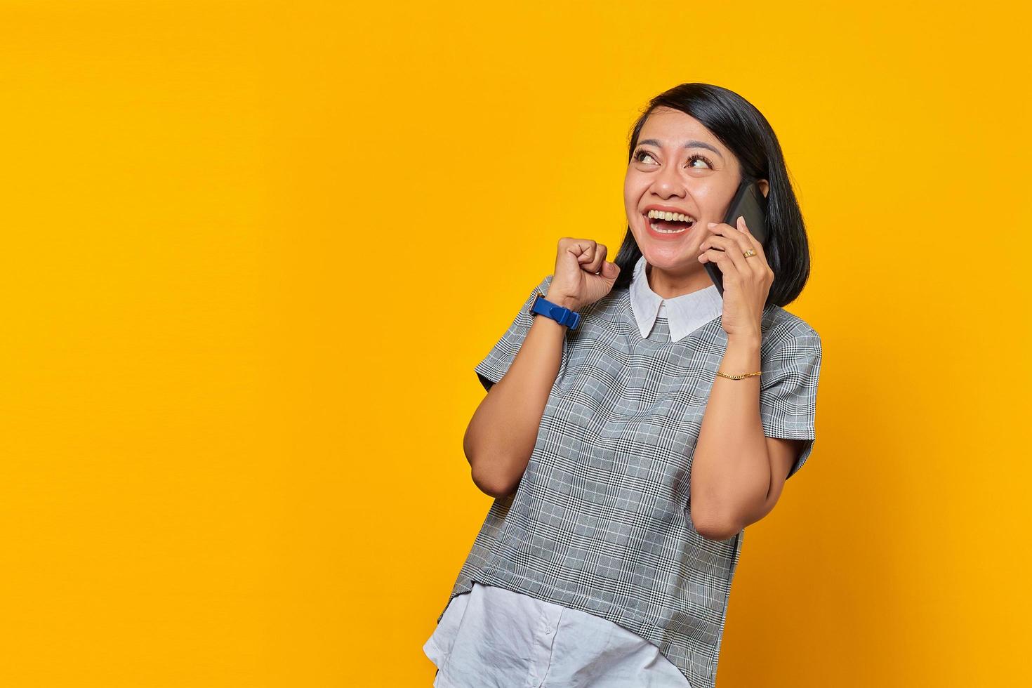 Excited young Asian woman receiving incoming call on smartphone and looking aside on yellow background photo