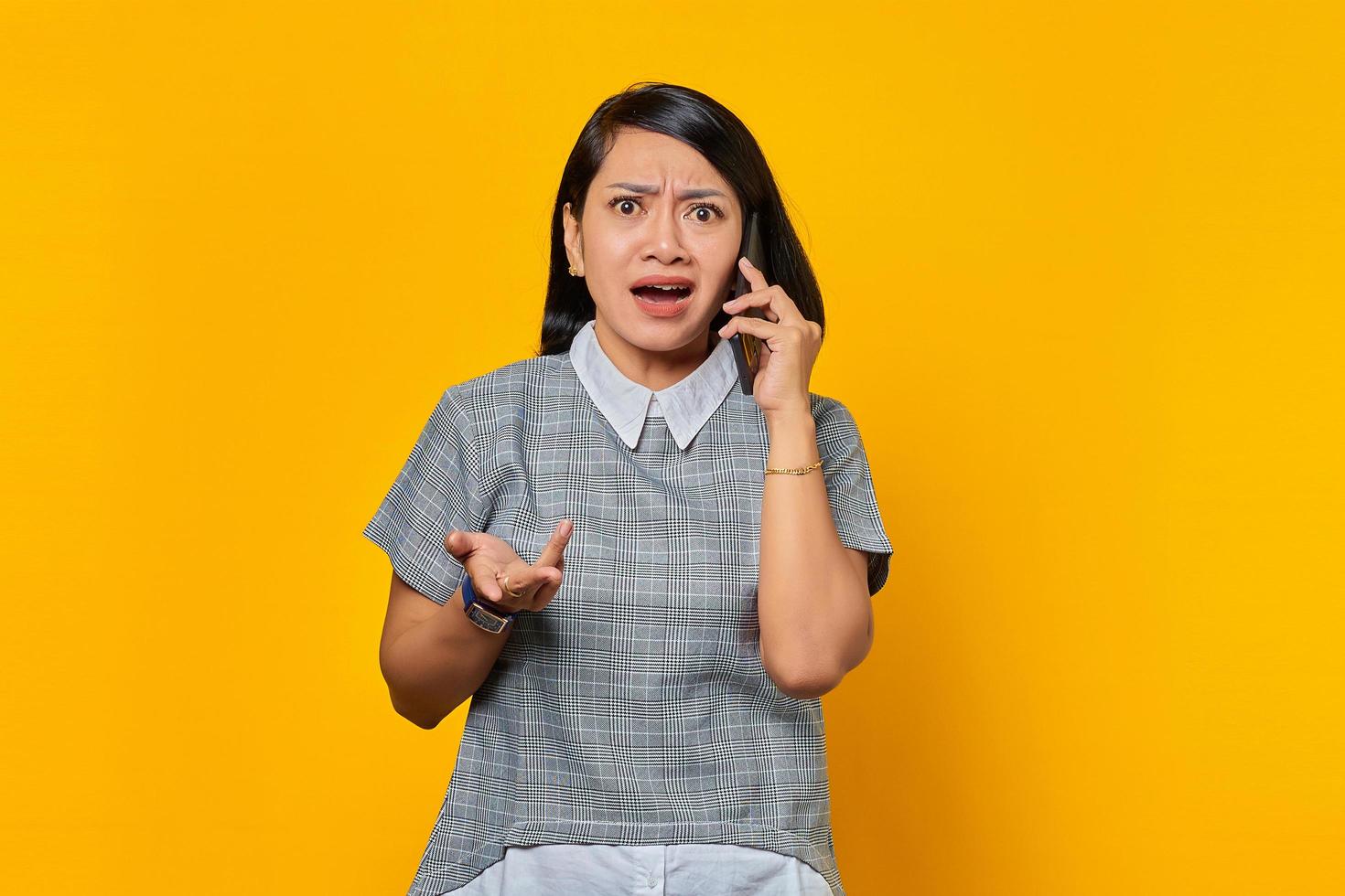 Portrait of angry young Asian woman talking on mobile phone with raised hands over yellow background photo