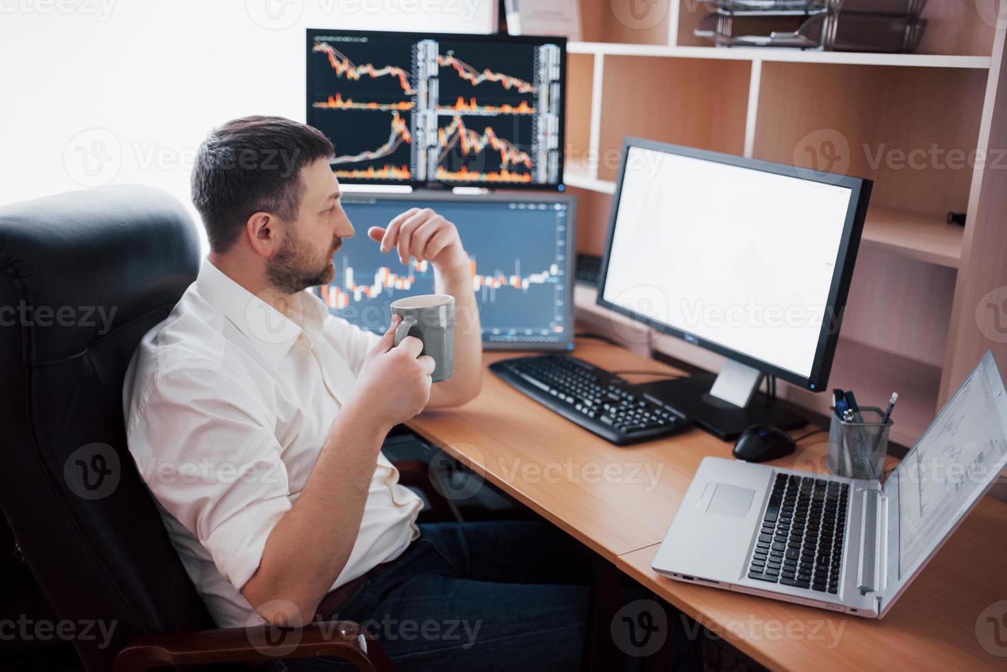 Young businessman is sitting in office at table, working on computer with many monitors,diagrams on monitor. Stock broker analyzes binary options charts.Hipster man drinking coffee,studying photo