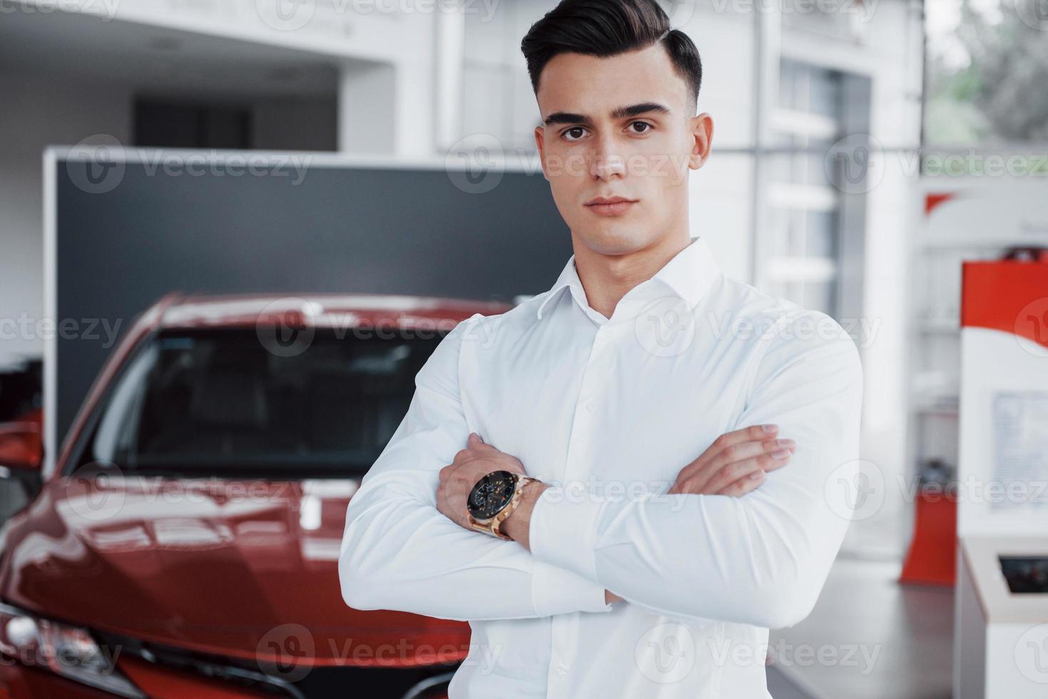 un hombre guapo es un comprador de pie junto a un coche nuevo en el centro de concesionarios y mirando a la cámara. foto