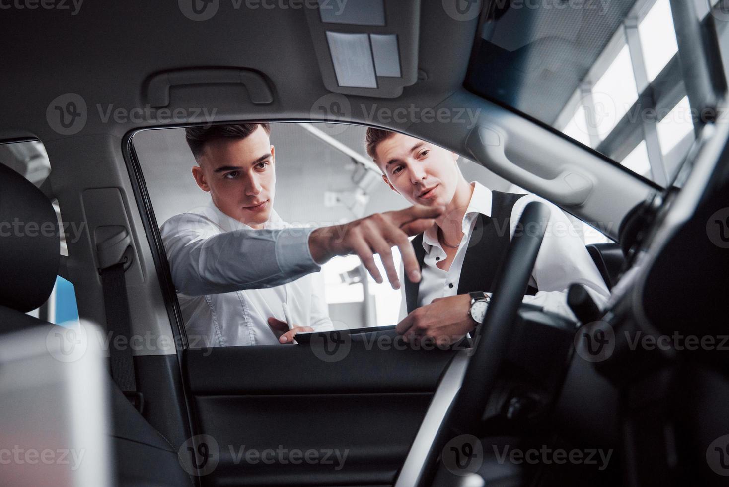 Two men stand in the showroom against cars. Close-up of a sales manager in a suit that sells a car to a customer. The seller gives the key to the customer. photo