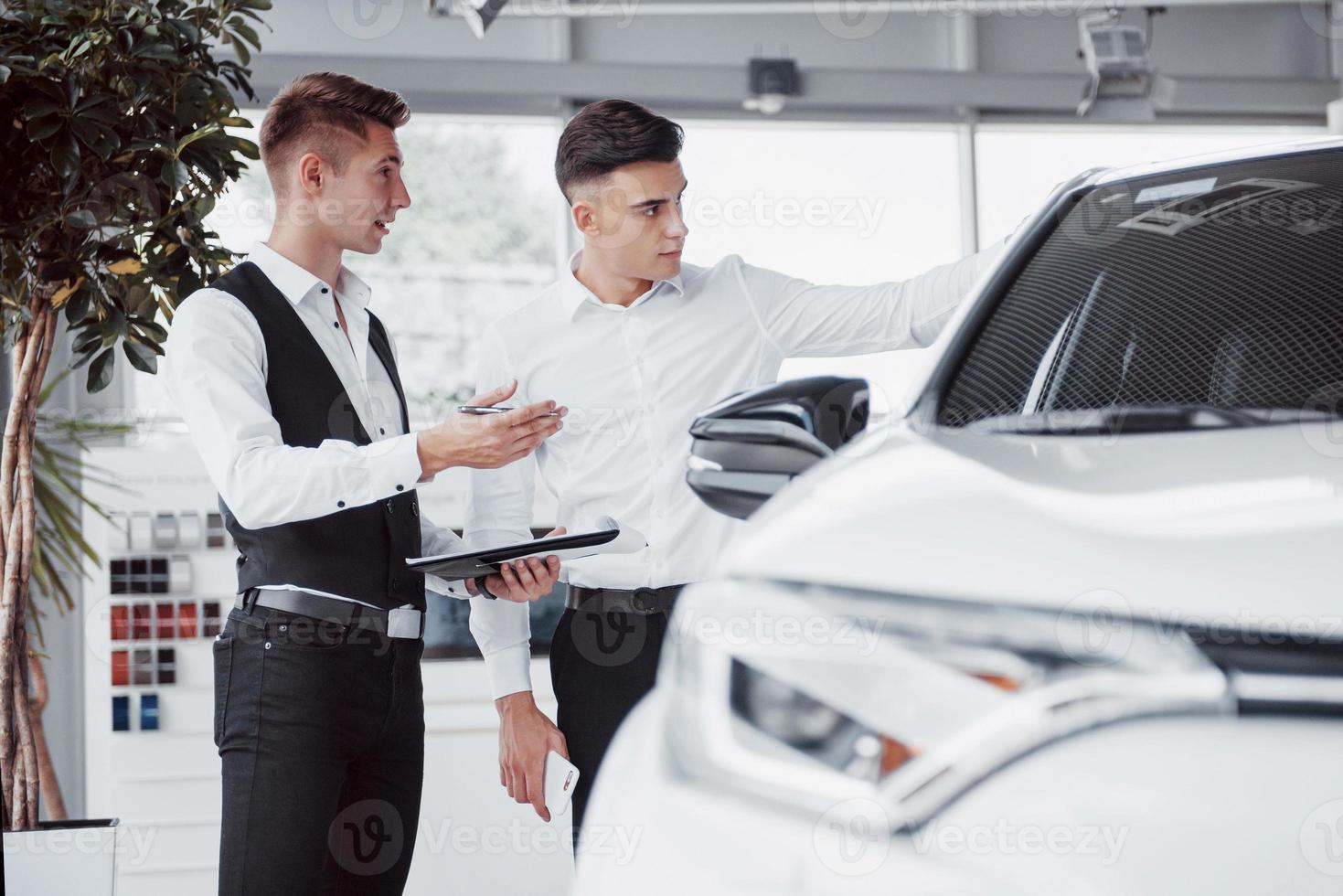 Two men stand in the showroom against cars. Close-up of a sales manager in a suit that sells a car to a customer. The seller gives the key to the customer. photo