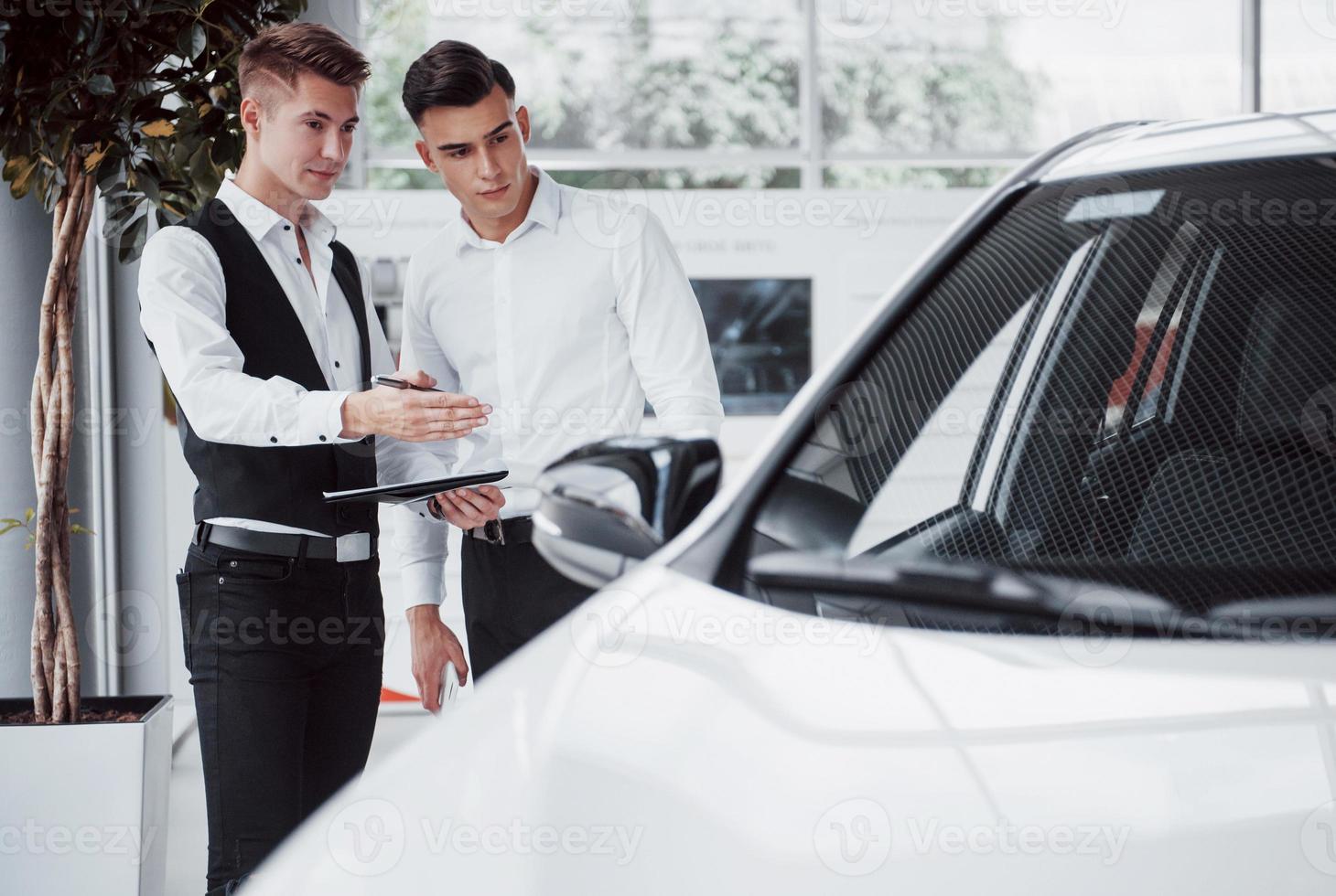 Two men stand in the showroom against cars. Close-up of a sales manager in a suit that sells a car to a customer. The seller gives the key to the customer. photo