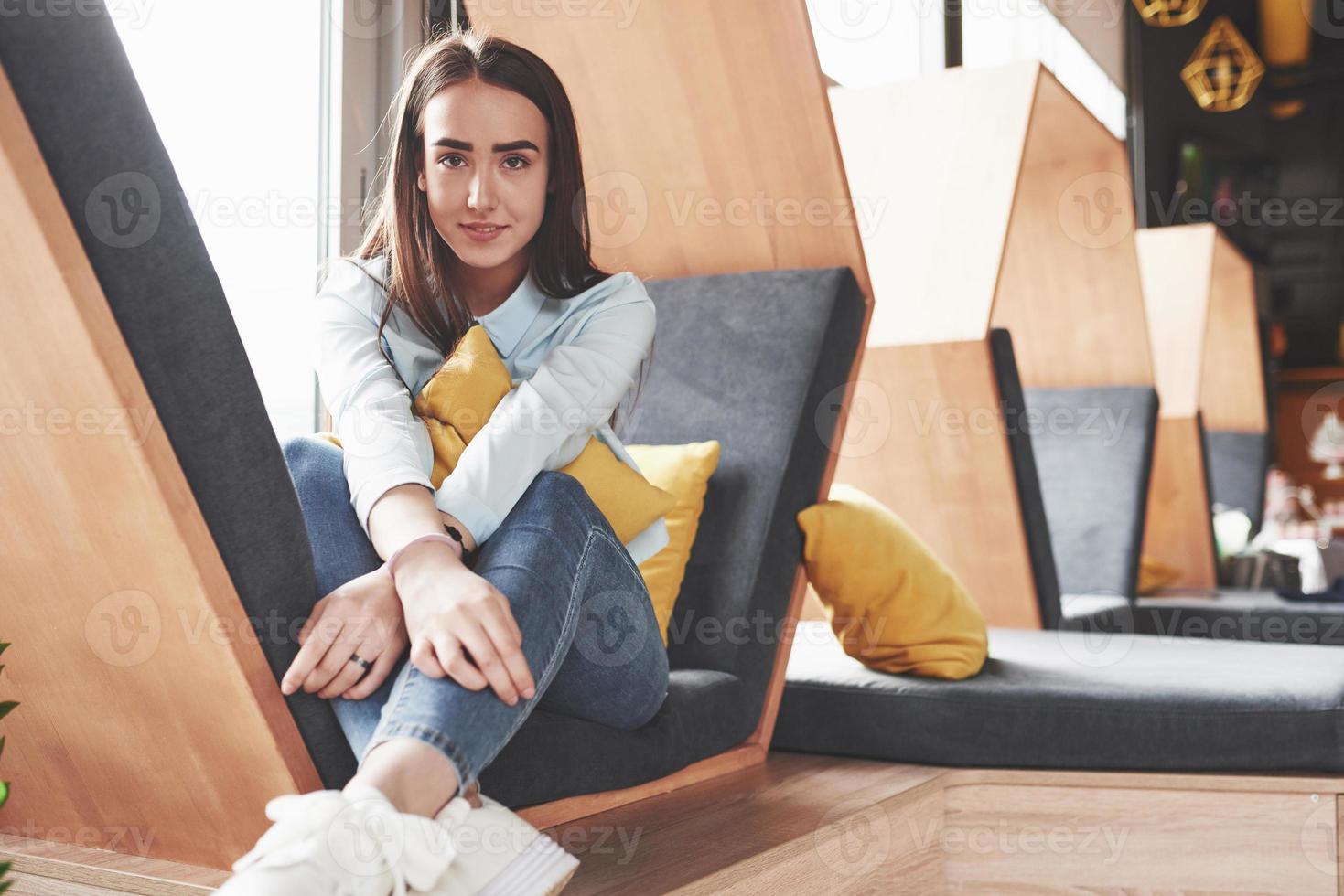 Beautiful young girl spends her time in the recreation area. She sits and has fun in the sofa chair in the form of a hexagon with pillows near the window photo