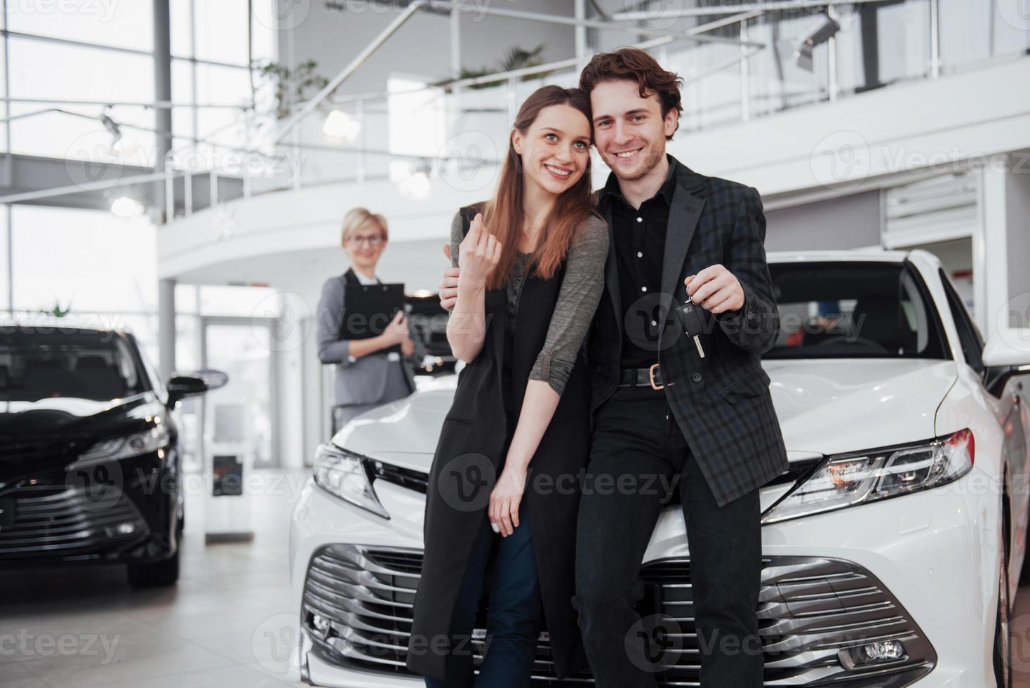 ahora su sueño se hace realidad. Dos felices jóvenes propietarios de coche atractivo, vendedor elegante en segundo plano. foto