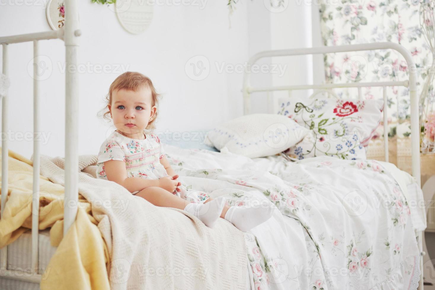 niña en vestido lindo emplazamiento en la cama jugando con juguetes en la casa. Childroom blanco vintage. concepto de infancia foto