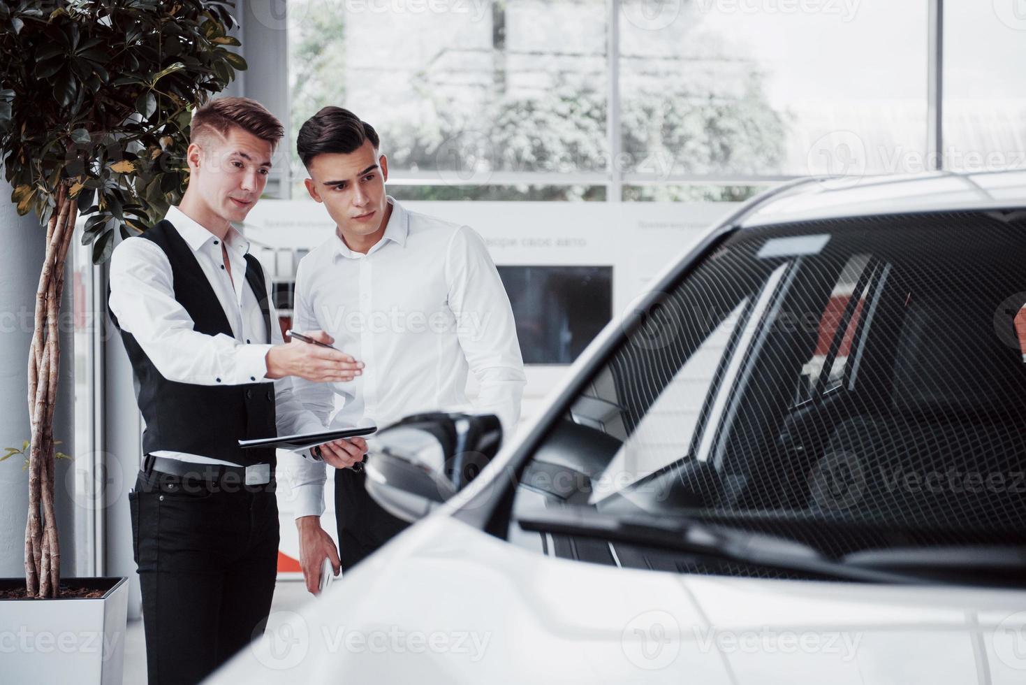 dos hombres se paran en la sala de exposición contra los coches. primer plano de un gerente de ventas con un traje que vende un automóvil a un cliente. el vendedor le da la llave al cliente. foto