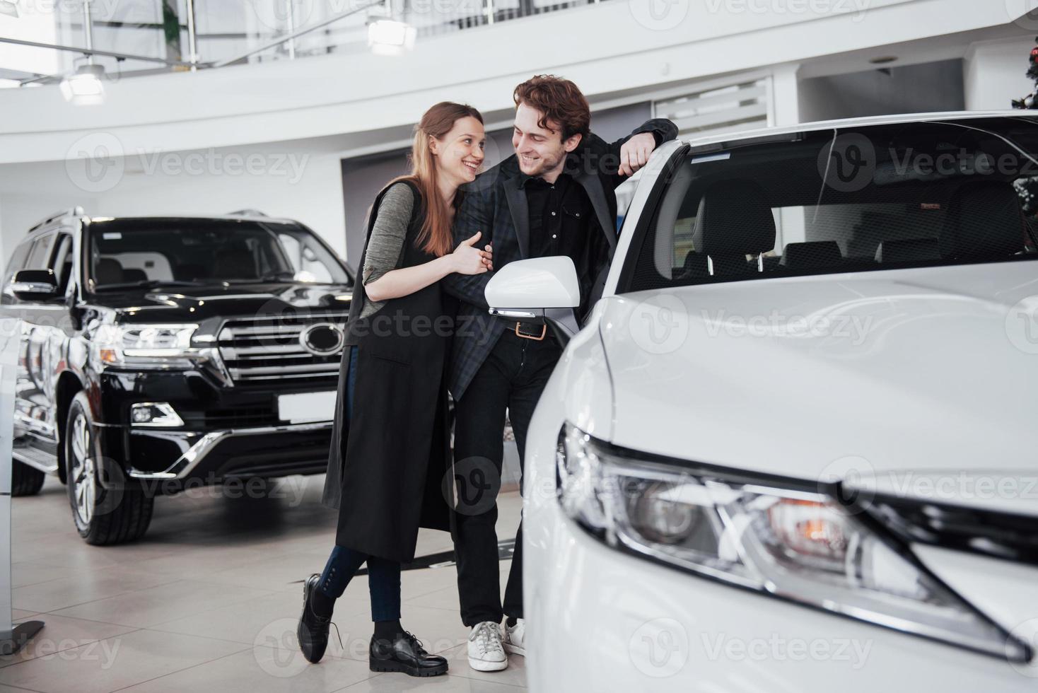 me gusta este coche. Hermosa joven pareja de pie en el concesionario eligiendo el coche para comprar foto