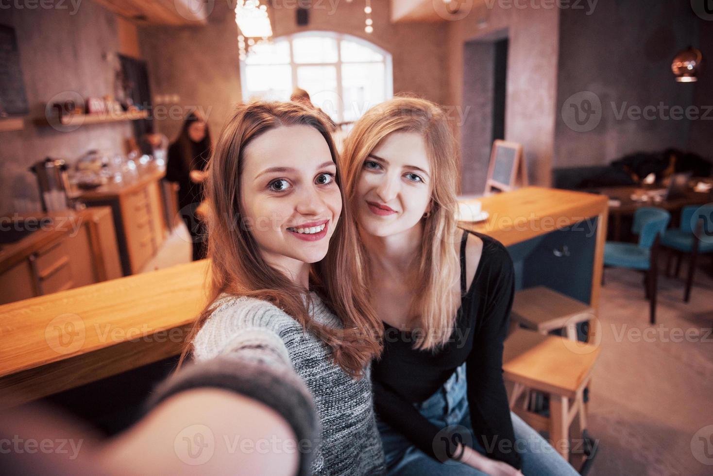 Two friends drinking coffee in a cafe, taking selfies with a smart phone and having fun making funny faces. Focus on the girl on the left photo