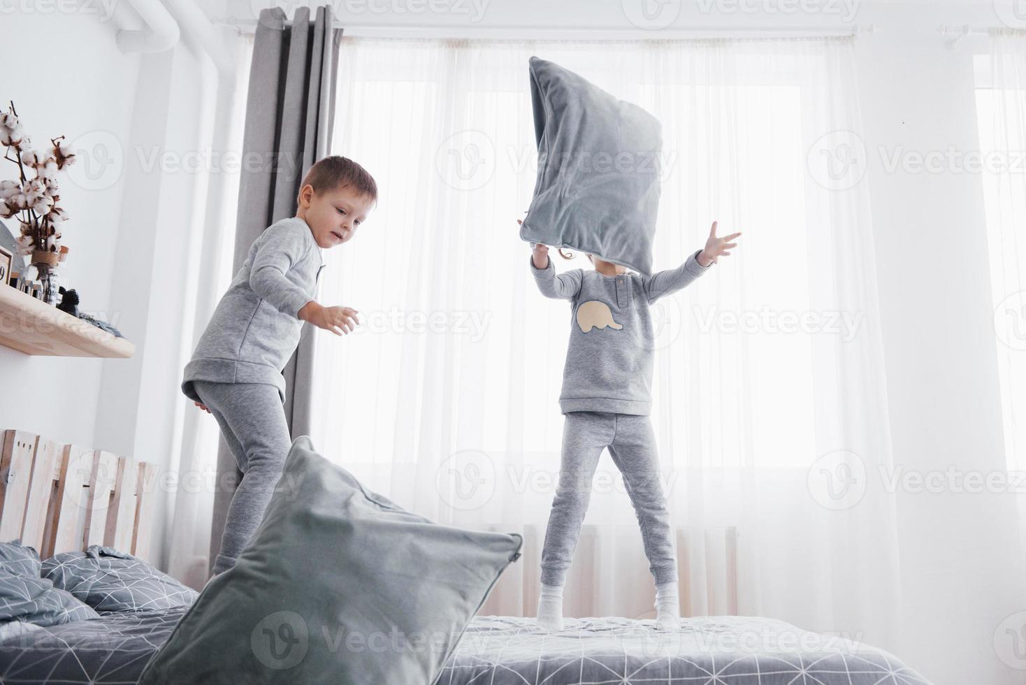 niños felices jugando en el dormitorio blanco. niño y niña, hermano y hermana juegan en la cama en pijama. ropa de dormir y ropa de cama para bebés y niños pequeños. familia en casa foto