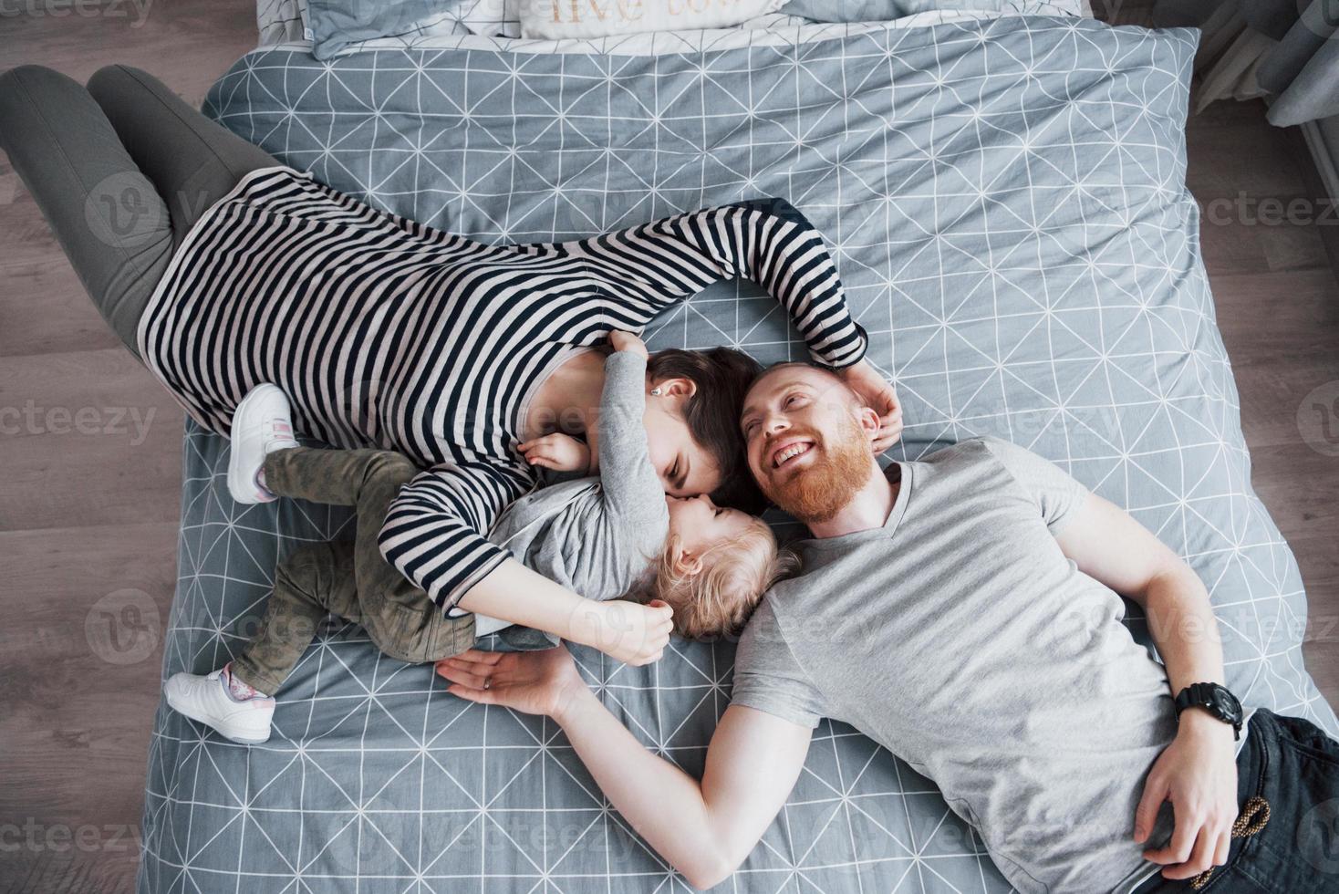 Vista superior de la hermosa joven madre, padre e hija mirando a la cámara y sonriendo mientras está acostado en la cama cabeza a cabeza foto